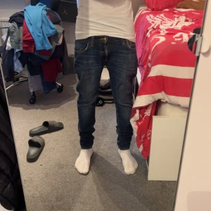 Blå jeans från Nudie Jeans - Säljer ett par klassiska blå jeans från Nudie Jeans med snygga slitningar och kontrastsömmar. Modellen har fem fickor, orange broderi på bakfickan och slim passform. Perfekta till vardags för en stilren look.w28 l30 nypris 1700!