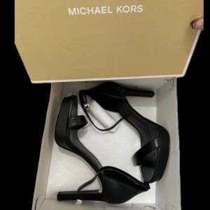 Svarta pumps från Michael Kors - Säljer ett par eleganta svarta pumps från Michael Kors med öppen tå och klack. Skorna har en stilren design med smala remmar och guldfärgad MK-detalj vid ankeln. Perfekta för dig som vill ha en klassisk och trendig look.