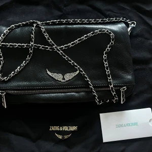 Svart axelväska från Zadig & Voltaire - Snygg svart axelväska i skinn från Zadig & Voltaire med silverfärgad kedjerem och dragkedja. Väskan har dekorativa vingar i metall framtill och flera fack med dragkedja. 