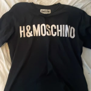 Svart t-shirt från H&MOSCHINO - Svart t-shirt från H&MOSCHINO med stor vit logga framtill. Klassisk rund halsringning och korta ärmar. Perfekt för dig som gillar streetstyle och vill sticka ut med en ikonisk logga. Hämtas i Västerhaninge tar endast kontanter 