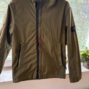 Olivgrön jacka från Stone Island Junior - Säljer en olivgrön jacka från Stone Island Junior med huva och dragkedja framtill. Jackan har två fickor med dragkedja och klassisk Stone Island-logga på ärmen. Perfekt för vår och höst. En liten söm som gått upp se bild. 