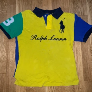Pikétröja från Ralph Lauren - Säljer en färgstark gul pikétröja från Ralph Lauren med blå krage och detaljer samt grön ärm med siffra. Stor logga och broderad text framtill. Klassisk piké med korta ärmar och knappar vid halsen.