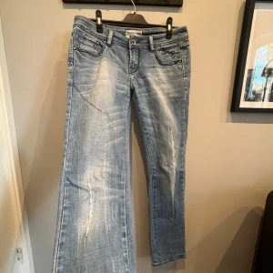 Ljusblå lågmidjade jeans med nitar. Köpta utomlands.  - Ljusblå jeansbyxor med slitningar och raka, vida ben. Dekorerade med små silverfärgade nitar på fram- och bakfickor samt broderad text på ena fickan. Klassisk femficksmodell med knapp och dragkedja.