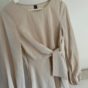 Beige blus - Säljer en stilren beige blus med lång ärm och snygg knytdetalj i midjan. Blusen har rund halsringning och en lätt utsvängd passform nedtill. Perfekt för dig som gillar enkla men trendiga plagg.