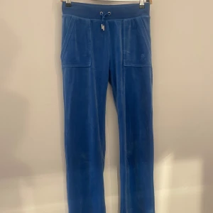 Blå velourbyxor från Juicy Couture - Mysiga blå velourbyxor från Juicy Couture med dragsko i midjan och stora fickor fram. Baktill finns två fickor med lock och knappar i guld. Perfekta för en avslappnad och trendig look.