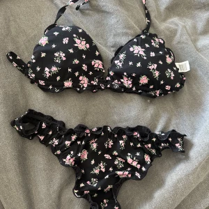 Svart blommig bikini med volang - Säljer en svart bikini med små rosa och vita blommor. Både överdel och underdel har volangdetaljer och bikinin har en feminin och söt stil. Överdelen har knyt i nacken och underdelen har rynkade kanter. ENDAST TESTAD!