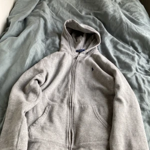 Grå hoodie från Polo Ralph Lauren - Säljer en klassisk grå hoodie från Polo Ralph Lauren med dragkedja och huva. Tröjan har två fickor framtill och den ikoniska lilla broderade loggan på bröstet. Perfekt för en avslappnad stil. Storlek 170. 
