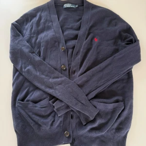 Mörkblå kofta från Polo Ralph Lauren - Mörkblå kofta med knappar framtill och två fickor. Klassisk design med röd broderad logga på bröstet. Perfekt för lager-på-lager och enkel att matcha med olika outfits.