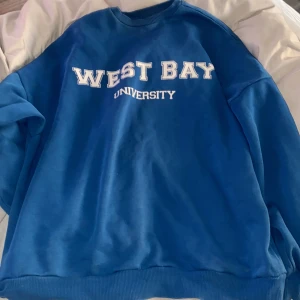 Blå sweatshirt med tryck  - Blå oversized sweatshirt med vit text 'West Bay University' på bröstet. Klassisk rund halsringning och ribbade muddar vid ärmslut och nederkant. Perfekt för en avslappnad och sportig stil.