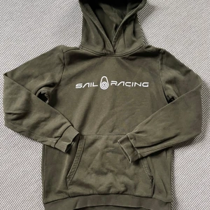 Sail Racing - Olivgrön hoodie - Säljer en olivgrön hoodie från Sail Racing med vit logga på bröstet och liten logga på huvan. Tröjan har magficka och ribbade muddar vid ärmslut och nederkant. Perfekt för en avslappnad stil. Storlek 150 (barn/ungdom). 