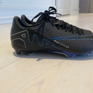 Svarta Nike fotbollsskor med blå detaljer - Säljer ett par svarta Nike fotbollsskor med blå och silvriga detaljer. Skorna har snörning och en platt sula med dobbar för bra grepp på planen. Swoosh-loggan syns tydligt på ovansidan och sidan. Perfekta för dig som vill ha stilrena och sportiga skor på matchen. 24cm långa på insidan