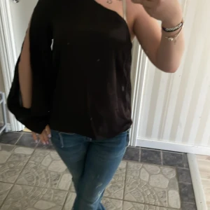 Svart oneshoulder blus - Säljer en svart oneshoulder blus med lång ärm och öppen axel från HM i storlek XS. Blusen har en snygg knappdetalj vid ärmslutet, säljer för att jag tycker inte att de är min stil