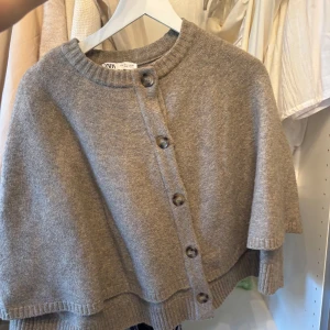 Beige stickad kofta från Zara - Mysig beige stickad kofta från Zara med rund halsringning och stora knappar framtill. Koftan har en lös passform och ribbade kanter vid hals och ärmslut. Perfekt för lager-på-lager under kyliga dagar.