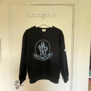 Svart sweatshirt från Moncler - Säljer en svart sweatshirt från Moncler med stort vitt logotryck på bröstet och rund hals. Tröjan har långa ärmar och klassisk passform. Perfekt för en stilren och avslappnad look.