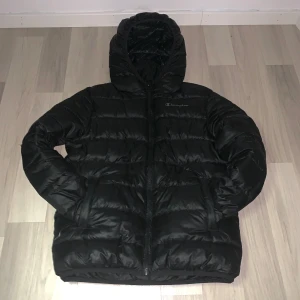 Svart pufferjacka från Champion - Säljer en svart pufferjacka från Champion med huva och dragkedja. Jackan har en klassisk pufferlook och är perfekt för kyligare dagar. Champion-loggan syns på bröstet. Snygg och enkel att matcha till det mesta. Den har endast en defekt i ryggen av jackan, det är ett litet hål