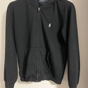 Svart hoodie med dragkedja från Polo Ralph Lauren - Svart hoodie från Polo Ralph Lauren med dragkedja framtill och huva. Klassisk logga broderad på bröstet och två fickor framtill. Modellen är custom fit och passar perfekt till en avslappnad stil. Storleken är S men passar också om du har M pris kan  sänkas vid snabb affär