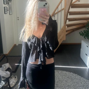 Svart blus med volang och knyt - Snygg svart blus med volangdetaljer och knyt framtill. Modellen är croppad och har långa ärmar med lätt utsvängda ärmslut. 