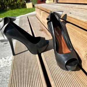 Svarta pumps med metallic rosett från ALDO - Säljer ett par svarta pumps från ALDO med hög klack och öppen tå. Skorna har en stor metallicfärgad rosett baktill som ger en unik och festlig look. Perfekta för dig som vill sticka ut med snygga detaljer.
