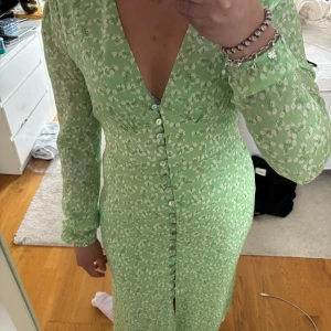 Ljusgrön Paris Dress från Adoore - Supersöt ljusgrön klänning från Adoore med små vita blommor. Djup v-ringning, långa ärmar och knappar hela vägen framtill. Figurnära passform och lätt glansigt material som känns lyxigt. Kapp sitter kvar på klänningen och den är helt oanvänd. Köparen står för frakten.