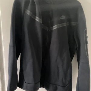 Nike Tech Fleece jacka svart och blå - Nike Tech Fleece jacka i svart och blå med dragkedja framtill och diskret Nike-logga på bröstet. Jackan har lång ärm och en modern, sportig look med svarta detaljer och ficka på ärmen. Byxor finns till den svarta tsm 1000kr men kan gå ner genom snabb affär 