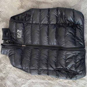 Svart dunväst från Emporio Armani EA7 - Säljer en svart dunväst från Emporio Armani EA7 med quiltad design och dragkedja framtill. Västen har en hög krage och två sidofickor med dragkedja. Perfekt för lager-på-lager och stilren look.