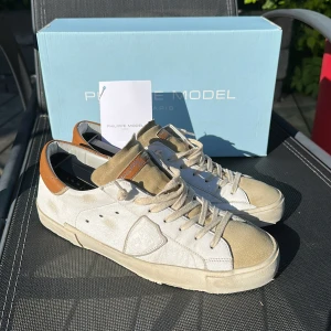 Philippe Model sneakers i beige och vitt - Säljer ett par Philippe Model sneakers i beige mocka och vitt skinn med bruna detaljer vid hälen. Skorna har snörning, rund tå och platt sula. Perfekta för dig som gillar stilrena och klassiska sneakers med en touch av lyx.