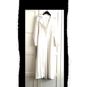 Vit långkappa med bälte från Suga - Säljer en elegant vit långkappa från Suga med lång ärm och tillhörande bälte i midjan. Kappan är tillverkad i polyester och har en stilren, minimalistisk design som passar perfekt till speciella tillfällen.