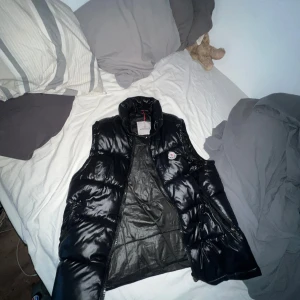 Svart dunväst från Moncler - Säljer en svart, glansig dunväst från Moncler med hög krage och dragkedja. Västen har Moncler-logga på bröstet och klassisk quiltad design. Perfekt för lager-på-lager och stilren look.