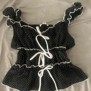 Prickig blus med volang och knyt - Söt svart blus med vita prickar, volangdetaljer och tre knytband framtill. Blusen har korta volangärmar och en figurnära passform. M/S