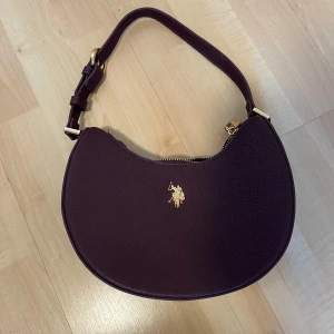 Mörklila axelväska från Ralph Lauren - Säljer en snygg mörklila axelväska från Ralph Lauren med guldfärgade detaljer och klassisk logga framtill. Väskan är tillverkad i skinn och har justerbart axelband samt dragkedja upptill. Perfekt accessoar för att lyfta din outfit.