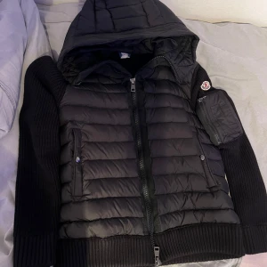 Svart pufferjacka med stickade ärmar från Moncler - Säljer en svart pufferjacka från Moncler med stickade ärmar och huva. Jackan har dragkedja framtill, två fickor med knappar och Moncler-logga på ärmen. Perfekt för kyliga dagar och riktigt snygg streetstil.