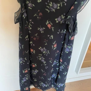 Blå blommig långklänning med volang - Blå klänning  med blommigt mönster i lila och rosa toner. Klänningen har volangdetaljer vid axlarna och är tillverkad i ett lätt, transparent material. Perfekt för dig som gillar romantisk stil och flowy känsla.