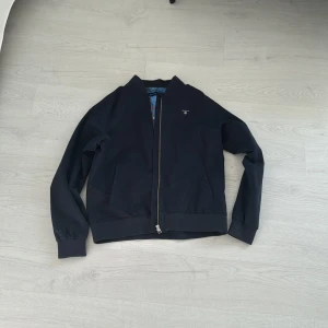 Svart bomberjacka från GANT - Snygg svart bomberjacka från GANT med klassisk passform och dragkedja framtill. Jackan har ribbade muddar vid ärmslut och midja samt två sidofickor. Fodret är blått med GANT-logga på insidan.