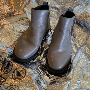 Glittriga boots med dragkedja - Säljer ett par snygga boots i glittrigt silverfärgat material. Skorna har rund tå, platt sula och dragkedja på sidan för enkel påtagning. Perfekta för att sticka ut lite extra!