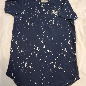 Blå t-shirt med vitt stänkmönster från SikSilk stl XS - Säljer en blå t-shirt från SikSilk med vitt stänkmönster och broderad logga på bröstet. T-shirten har rund hals och korta ärmar, perfekt för en avslappnad stil.