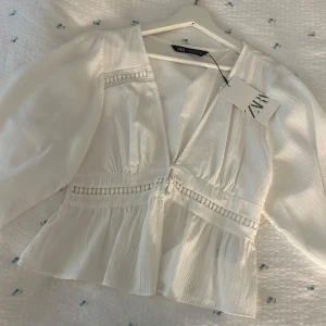 Vit blus med volang från Zara - Säljer en vit blus från Zara med v-ringning och volang nertill. Blusen har långa ärmar och dekorativa band med små bollar längs bysten och midjan. Tunt, lätt material som ger en luftig känsla. Aldrig använd prislapp kvar!
