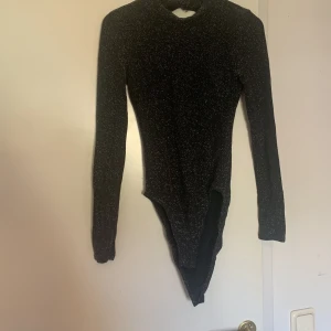 Svart glittrig body från H&M Divided - Svart glittrig body med lång ärm och hög hals från H&M Divided. Bodyn har en snygg öppen rygg och är figurnära i modellen. Perfekt för att sticka ut med lite extra glitter.