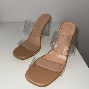 Beige klackar med transparent rem - Säljer ett par snygga beige klackar med transparenta remmar. Skorna har fyrkantig tå och öppen design, perfekta för att lyfta din outfit med en modern touch.