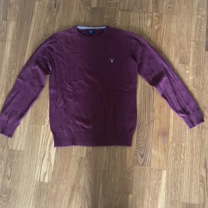 Vinröd tröja från GANT - Vinröd långärmad tröja från GANT med rund halsringning och liten broderad logga på bröstet. Tröjan har ribbade muddar vid ärmslut och nederkant. Perfekt för en stilren och klassisk look.