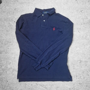 Marinblå pikétröja från Polo Ralph Lauren - Säljer en marinblå långärmad pikétröja från Polo Ralph Lauren med klassisk krage och knappar framtill. Tröjan har den ikoniska röda logotypen broderad på bröstet och är i en slim fit-modell. Perfekt för en stilren och tidlös look.