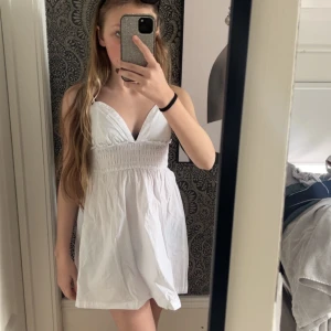 Vit kortklänning med v-ringning - Söt vit kortklänning med djup v-ringning och smockad midja. Klänningen är ärmlös och har en luftig passform, perfekt för varma dagar. Enkel och stilren design som passar till många tillfällen.