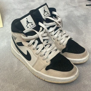 Nike Air Jordan 1 Mid Barely Orange  - Säljer ett par Nike Air Jordan 1 Mid i svart och beige med klassisk Jordan-logga på sidan och tungan. Skorna har högt skaft, svart swoosh och vita skosnören. Perfekta för dig som gillar streetwear och sneakers med ikonisk design. Materialet är mjukt tyg där av att det beiga blivit missfärgat lätt. Köpta för 4 år sedan. 