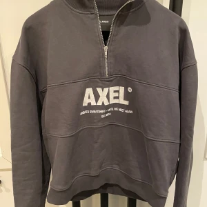 Grå sweatshirt med dragkedja från Axel Arigato - Grå sweatshirt med halv dragkedja och hög krage från Axel Arigato. Tryckt vit logga och text på bröstet. Tröjan har långa ärmar och en avslappnad passform, perfekt för en stilren vardagslook.