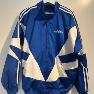 Blå och vit Adidas vindjacka - Retroinspirerad blå och vit vindjacka från Adidas med klassiska ränder på axlarna och grafiskt mönster. Jackan har dragkedja framtill, hög krage och ribbade muddar. Perfekt för en sportig look.