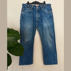 Levis 501 blå jeans med rå kant - Klassiska blå Levis 501 jeans med raka ben och råa, fransiga benslut. Jeansen har knäppning med knappar och naturlig sliten look. Snygga för en avslappnad och bekväm look. Jeansen har lite markeringar på framsida höger lår, där två figurer målats, samt två svaga rosa färgfläckar.