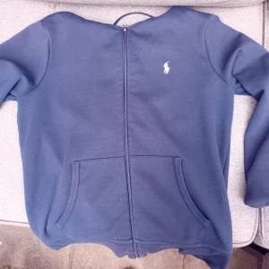 Kofta från Polo Ralph Lauren - Säljer en marinblå hoodie från Polo Ralph Lauren med dragkedja framtill och klassisk logga broderad på bröstet. Tröjan har långa ärmar och två fickor framtill. Perfekt för en avslappnad stil. Storlek M och 175cm. Priset är förhandlings bart