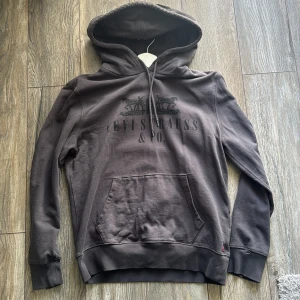 Mörkgrå hoodie från Levi's - Säljer en mörkgrå hoodie från Levi's med tryck framtill och klassisk känguruficka. Tröjan har huva med dragsko och ribbade muddar vid ärmslut och nederkant. Perfekt för en avslappnad stil.