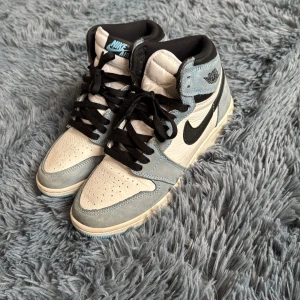 Nike Air Jordan 1 i blå och vitt - Säljer ett par Nike Air Jordan 1 med blå mockadetaljer, vit läderbas och svarta snören. Skorna har den klassiska Air Jordan-loggan på sidan och svart swoosh. Perfekta för dig som gillar streetwear och sneakers med stil. Knappast andvända, köpt för ca 5 tusen för 2-3 år sedan men sänker brutalt för jag inte vill ha dom nå mer. 39 men passar 40-41. Box och blåa Nike original skosnören ingår för extra 20kr. Kanske går att pruta😉