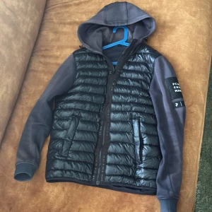 Svart hybridjacka med huva från Peak Performance - Säljer en svart hybridjacka från Peak Performance med vadderad framsida och ärmar samt rygg i sweatshirtmaterial. Jackan har huva, dragkedja och två fickor fram. Märkeslogga på ärmen. Har ett hål i luven men inget som syns vid användning och ett litet hål nere vid midjan, ser ni på bild 3,4. Den är solblekt men själv tycker jag de inte är så stor skillnad