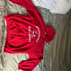Röd hoodie med Mackensen Bulldogs-tryck - Röd hoodie med vit text och tryck på bröstet där det står 'Mackensen Bulldogs' och en bulldog-illustration. Klassisk känguruficka framtill och huva. Perfekt för dig som gillar sportig stil.